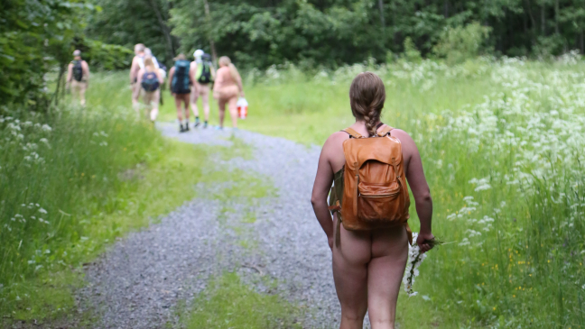 En naken person med ryggsekk går litt bakenfor en flokk med nakne turgåere i en lysning der stien grenser til en blomstereng.